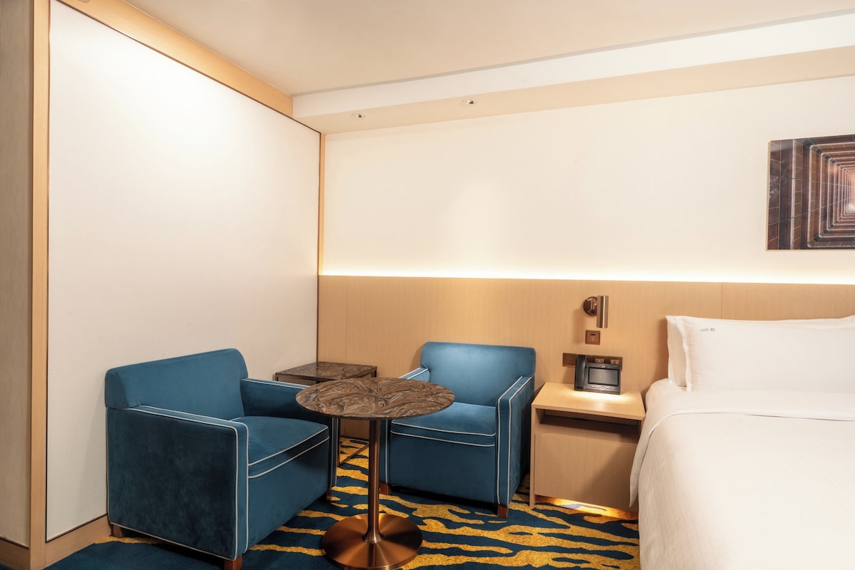 A hotel room with a bed, two blue armchairs, a small round table, a nightstand with a radio, and a patterned rug.