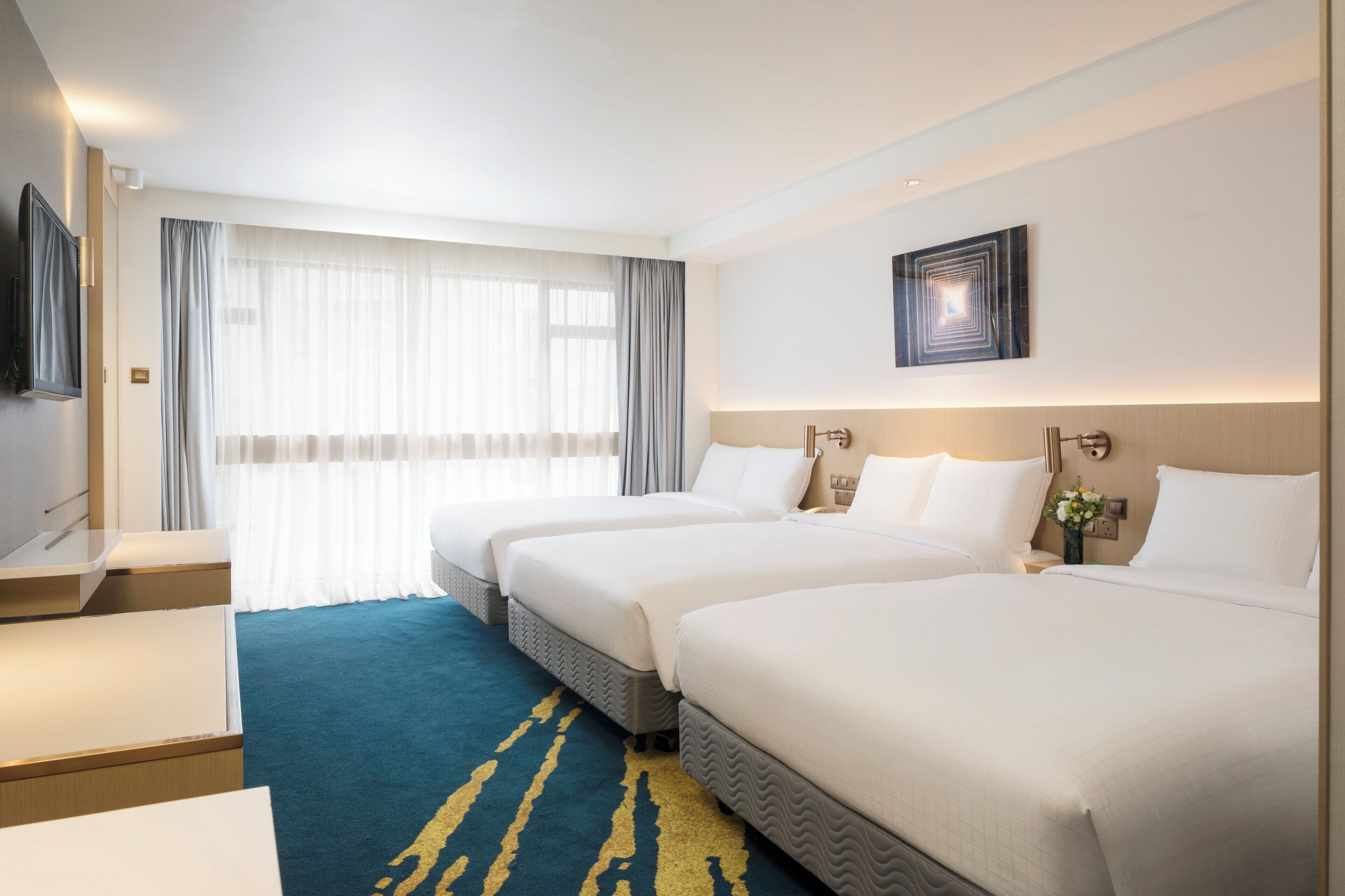 A hotel room with two beds, a desk, a TV, and a large window with curtains.