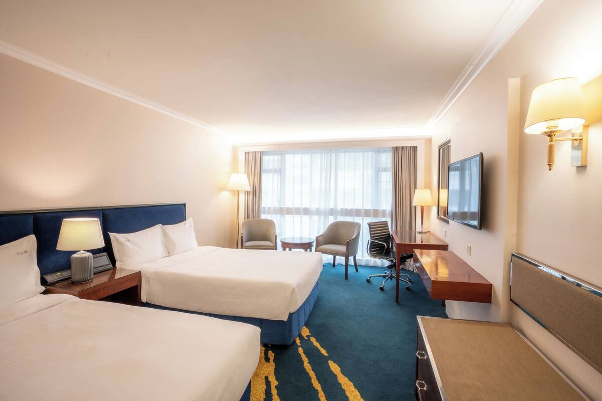 A hotel room with two beds, a desk, a chair, a TV, and a window with curtains.