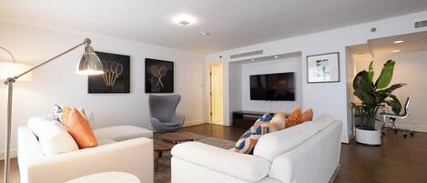 Ein modernes Wohnzimmer mit einem weißen Sofa, einem grauen Sessel, einem kleinen runden Tisch und einer Stehlampe. An der Wand ist ein Flachbildfernseher angebracht, es gibt einen Blumentopf und ein gerahmtes Bild.
