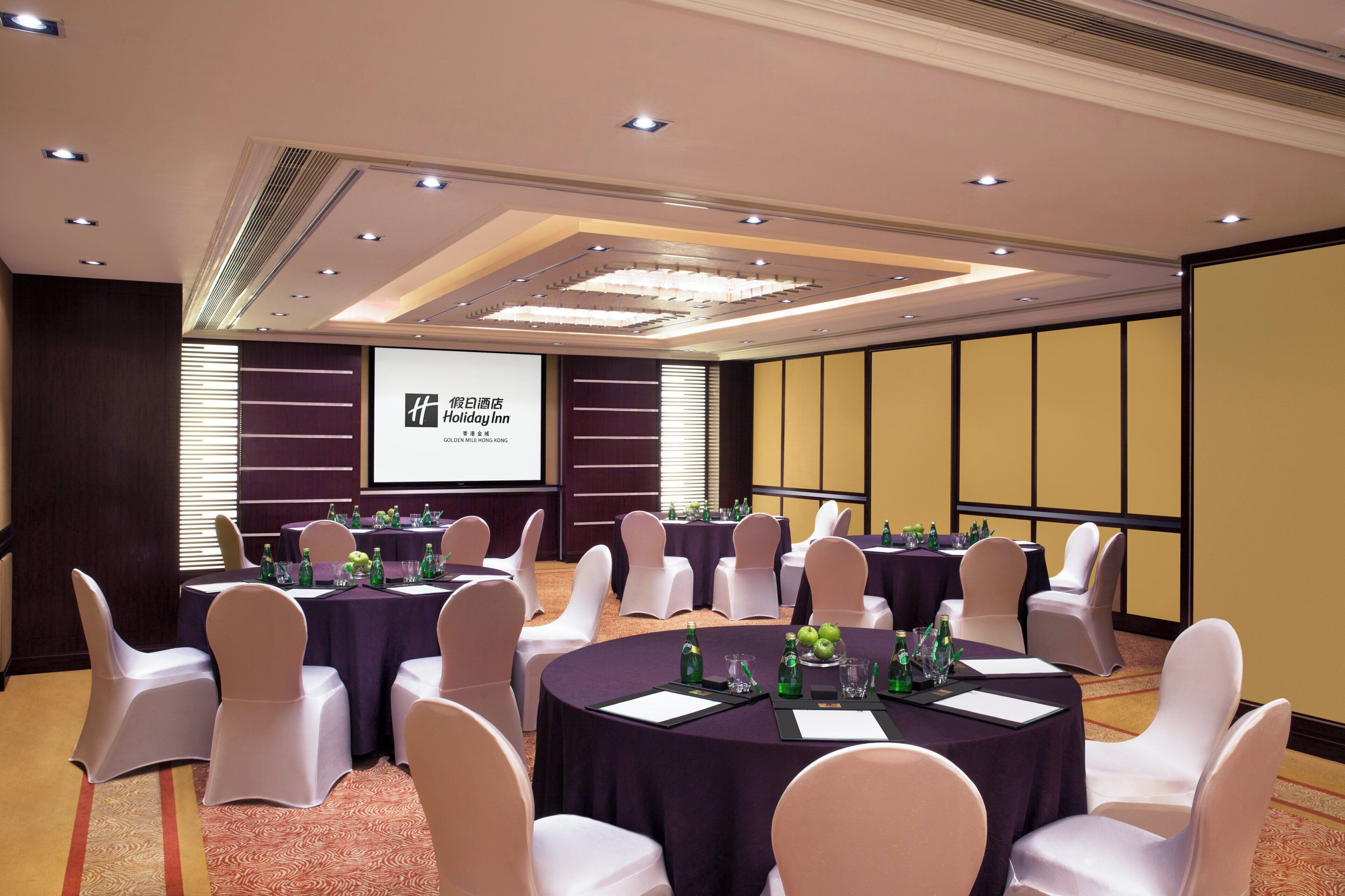 A conference room with round tables, white chairs, and a projection screen displaying 'Holiday Inn' branding.