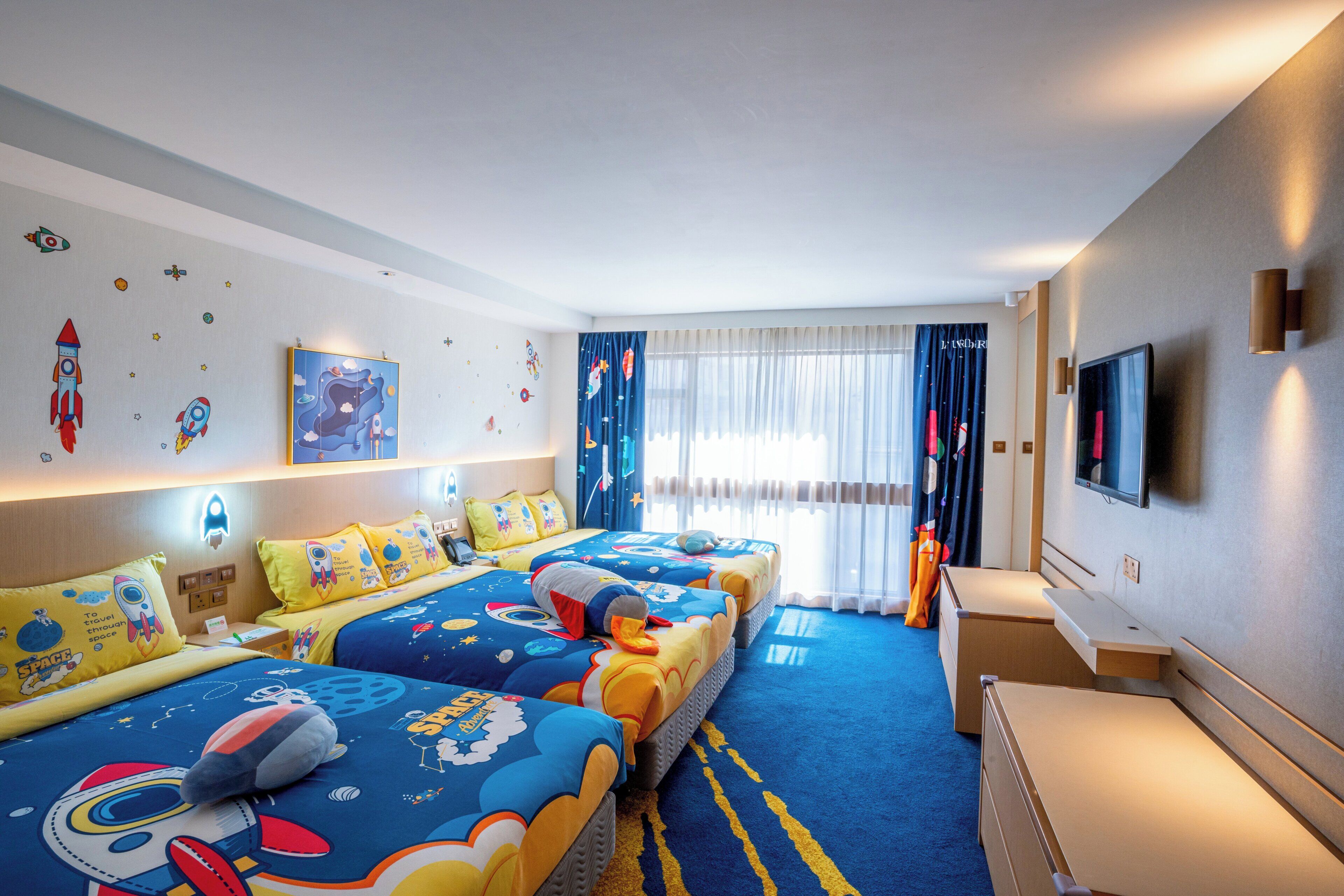 A hotel room with a large bed, a desk, a TV, and space-themed decor.