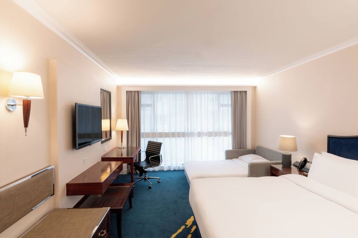 A hotel room with a bed, a desk with a chair, a TV, and a window with curtains.