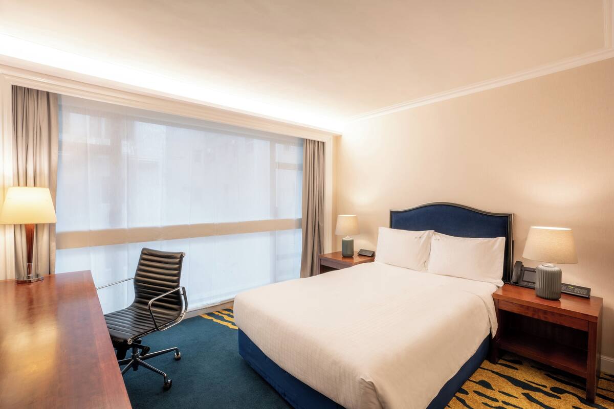 A hotel room with a bed, a desk, a chair, a nightstand, and a window with curtains.