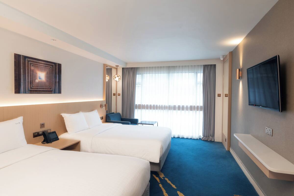 A hotel room with two beds, a desk with a phone, a TV mounted on the wall, and a large window with curtains.