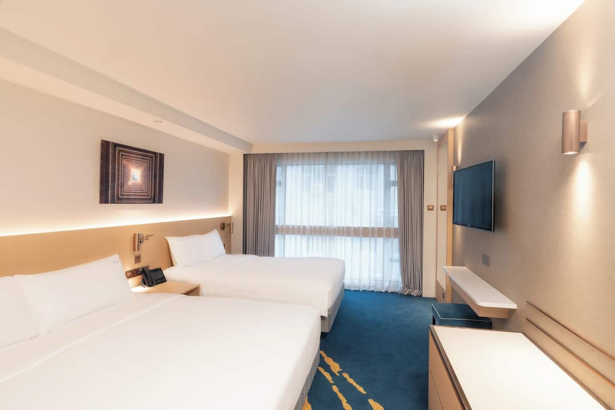 A hotel room with two beds, a desk, a TV, and a large window with curtains.