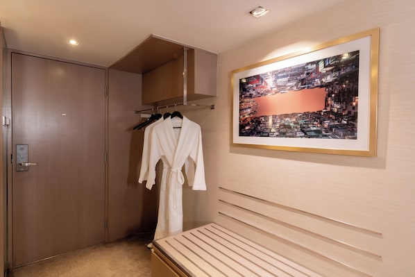 Une chambre d’hôtel avec une armoire en bois, une robe blanche suspendue, un lit avec une tête de lit et un tableau représentant un paysage urbain encadré accroché au mur.