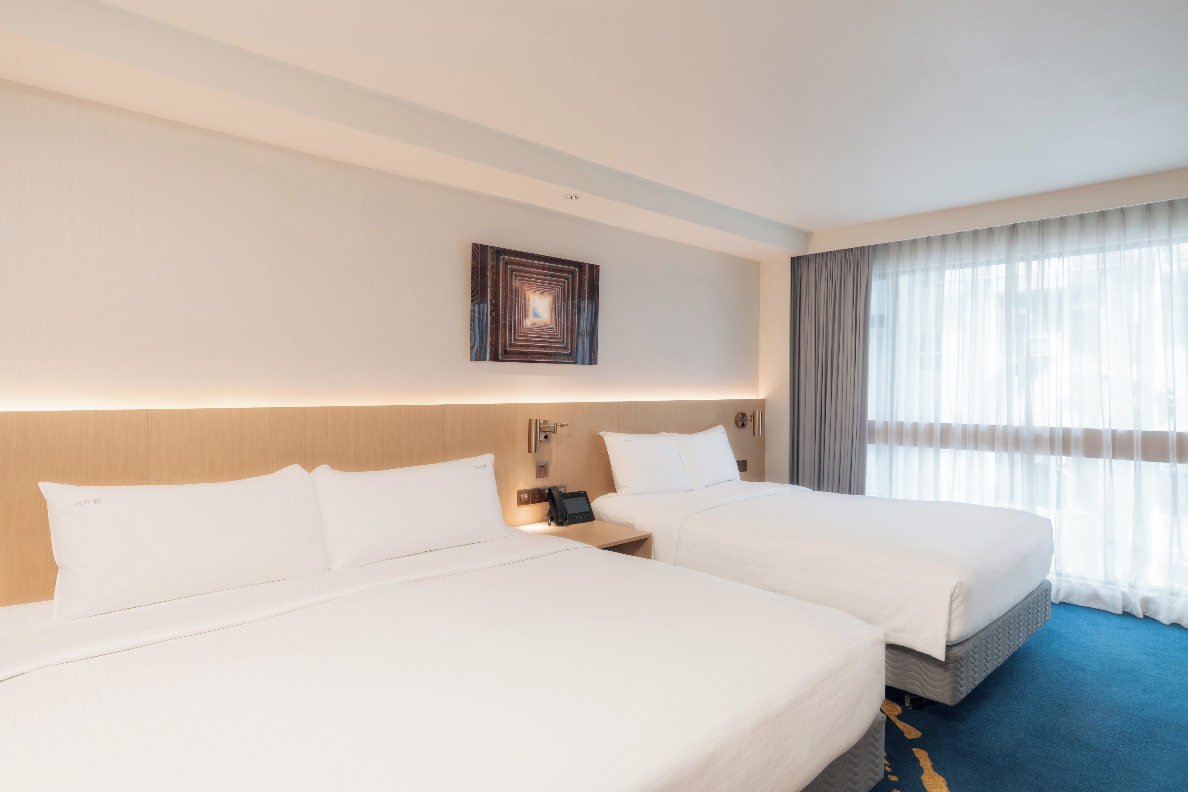 A hotel room with two beds, a bedside table with a phone, a wall-mounted artwork, and a large window with curtains.