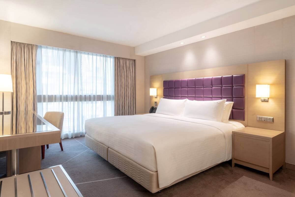 A hotel room with a large bed, a desk, and a chair. There is a window with curtains, a nightstand with a lamp, and a wall-mounted light.