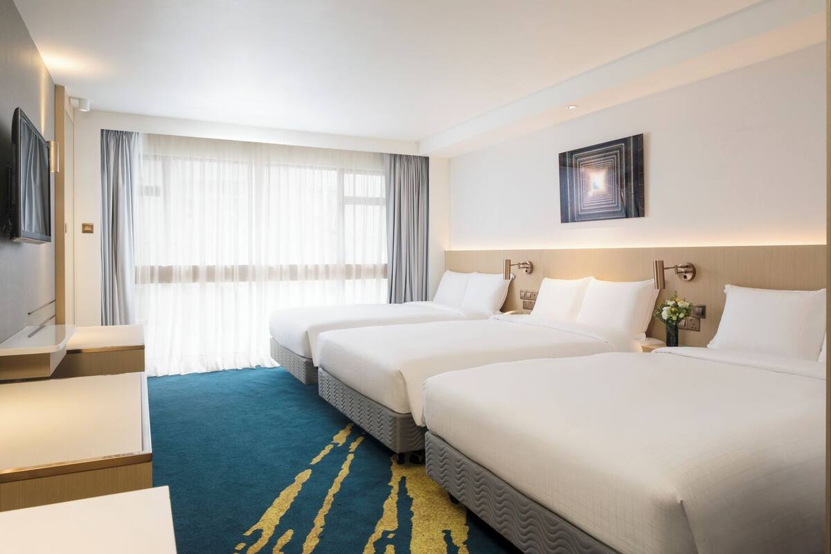A hotel room with two beds, a desk, a TV, and a large window with curtains.