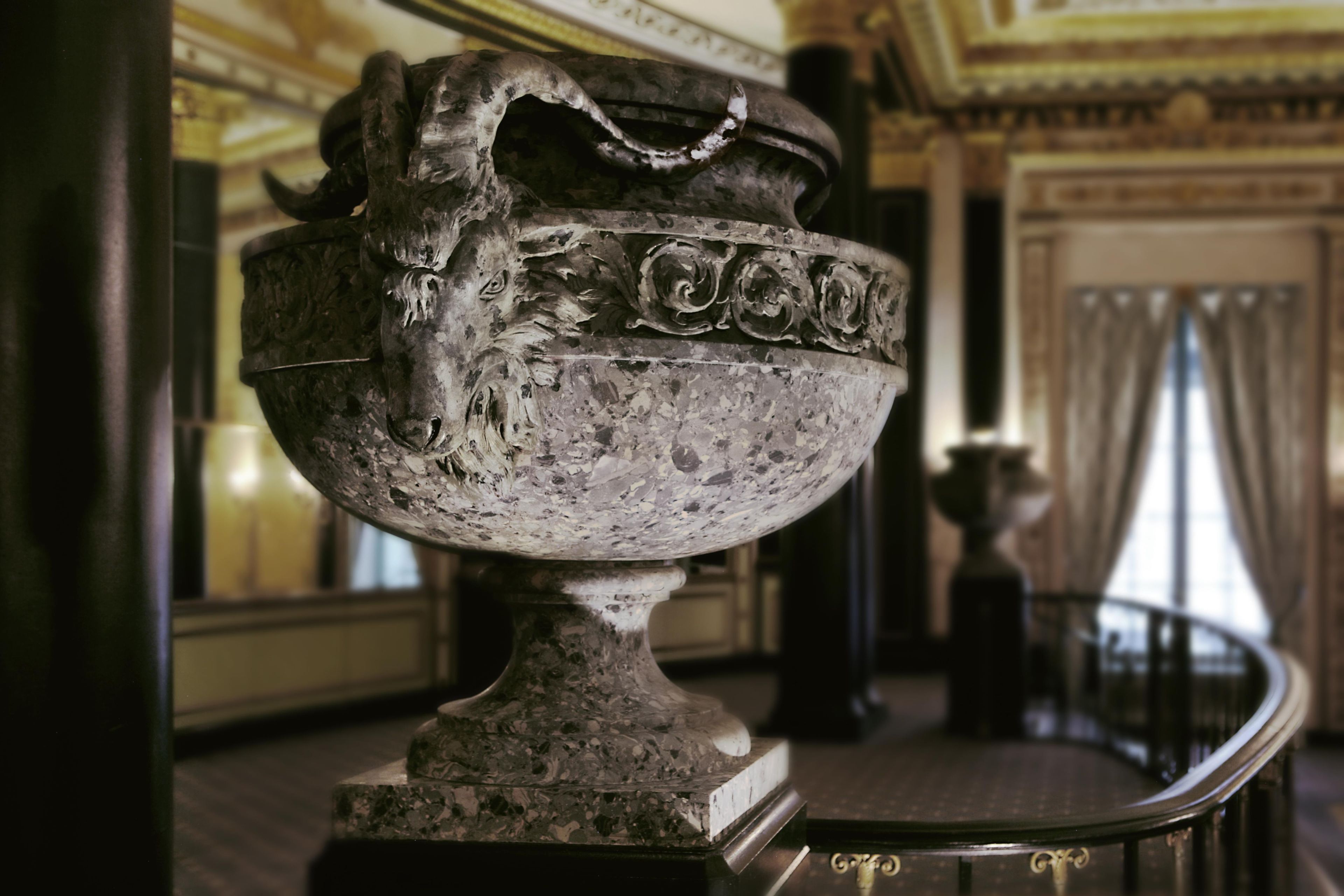 Un recipiente metálico grande y ornamentado con diseños intrincados, ubicado sobre un pedestal en una habitación de arquitectura clásica.