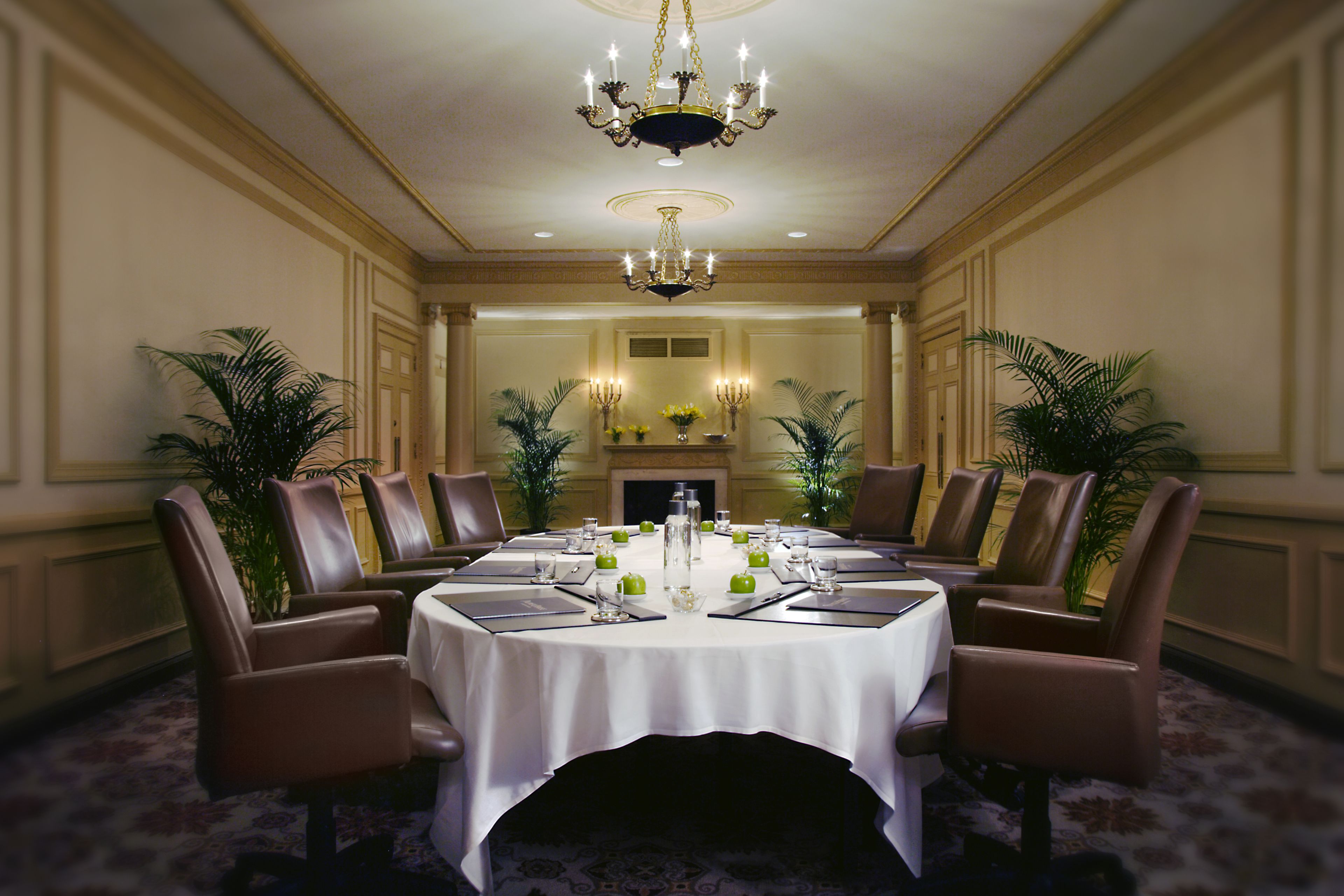 Un comedor formal con una mesa redonda dispuesta para ocho personas, rodeada de sillas de cuero, una araña y plantas en macetas.