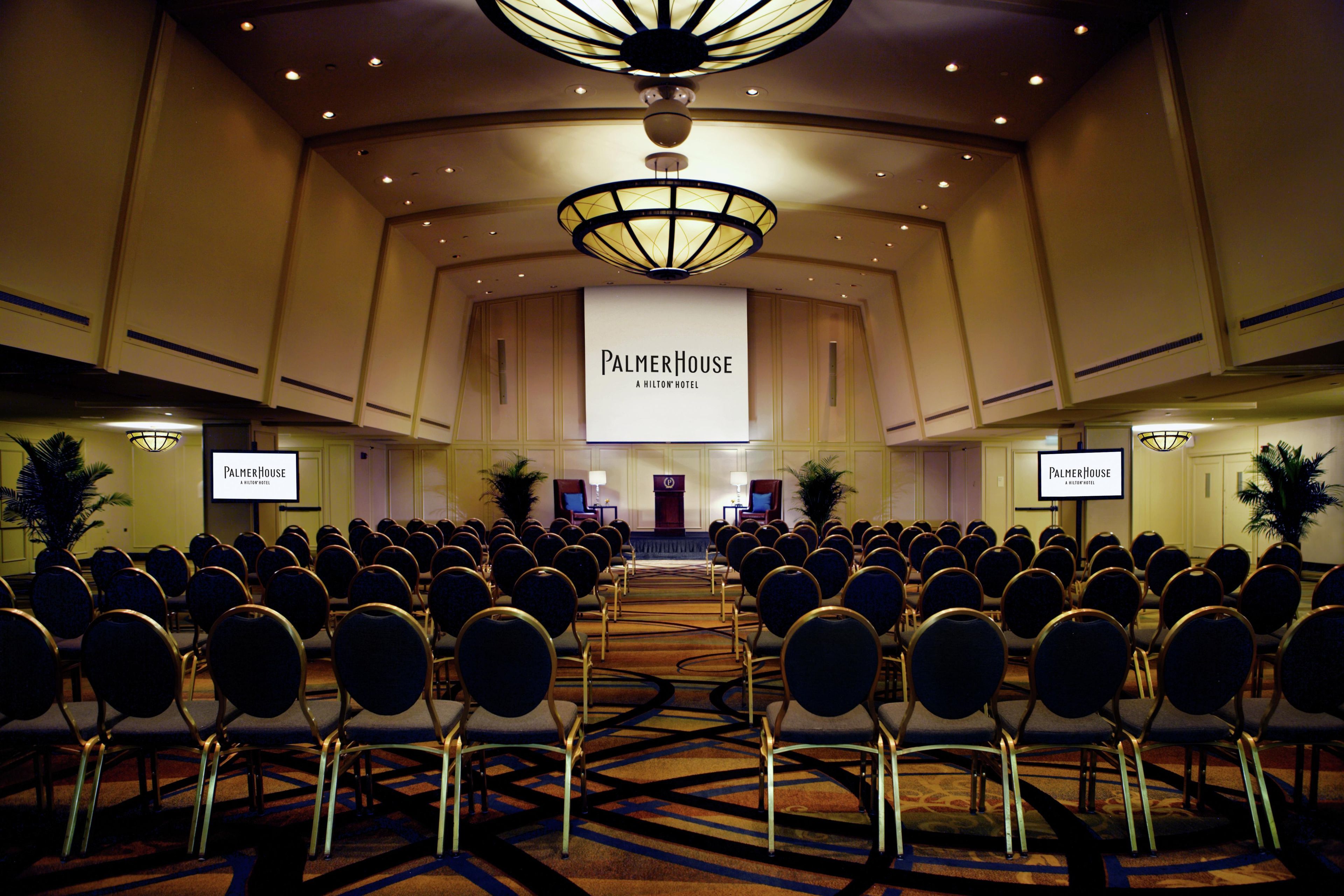 Een congreszaal met rijen stoelen, een spreekgestalte en een groot scherm waarop 'PalmerHouse A Hilton Hotel' staat.