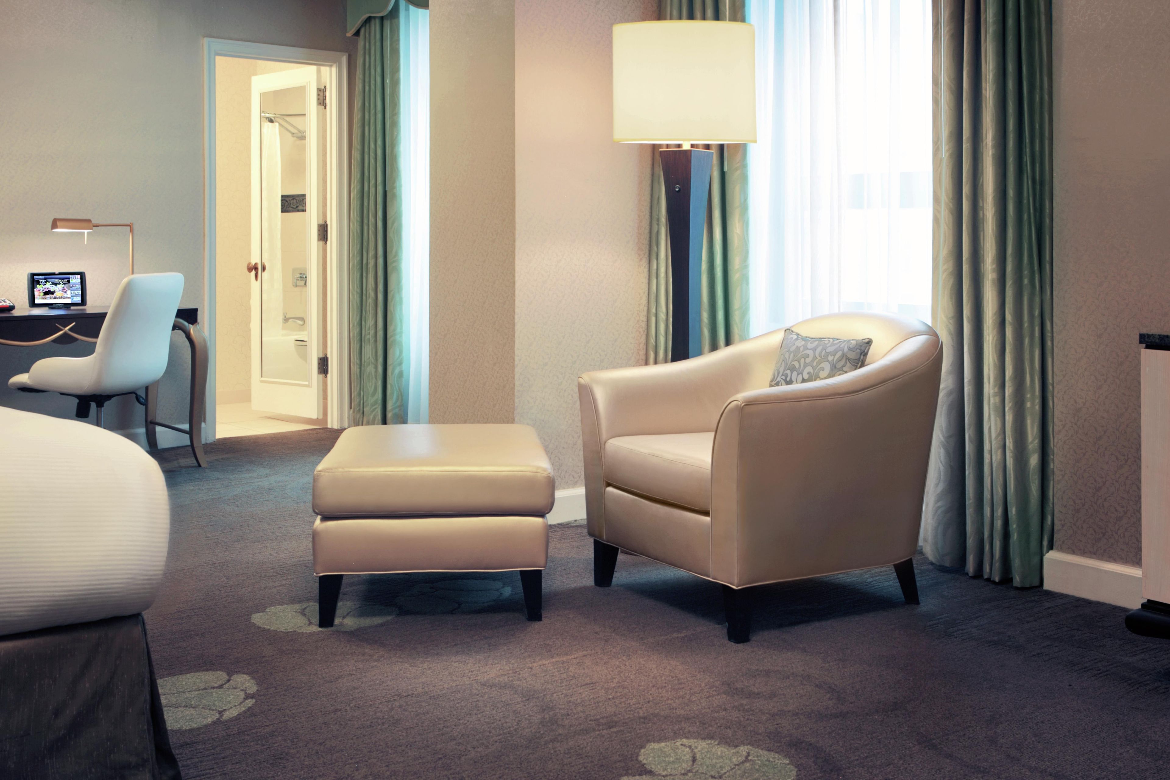 Una habitación de hotel con una cama, un escritorio con computadora, una silla, un reposapiés, una lámpara y una ventana con cortinas.