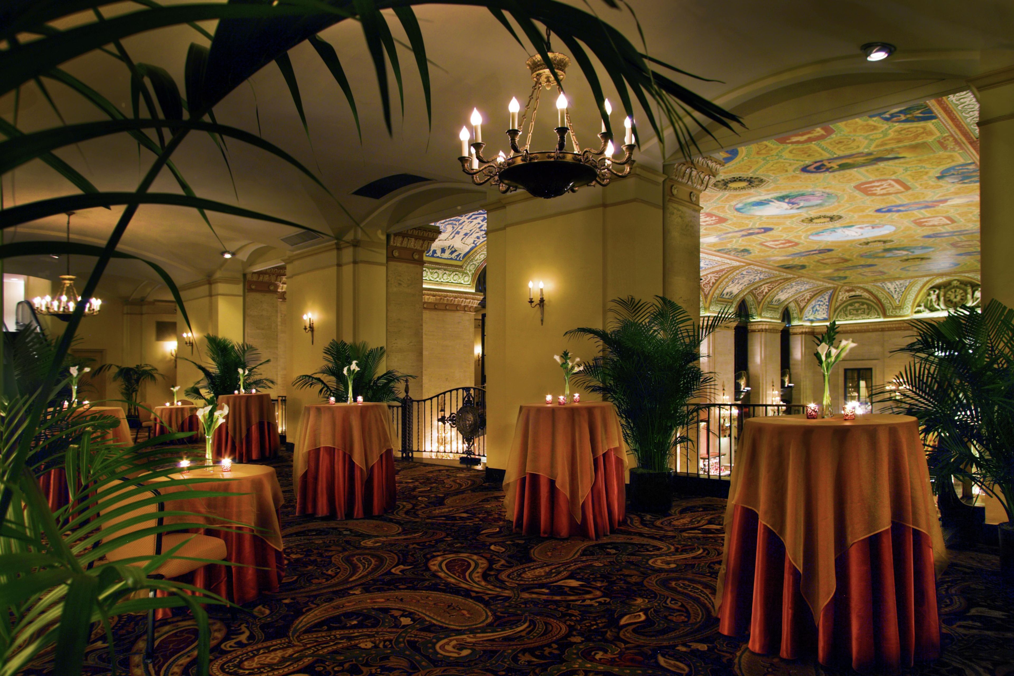 A hotel lobby with round tables covered in red tablecloths, a chandelier, and a patterned carpet.