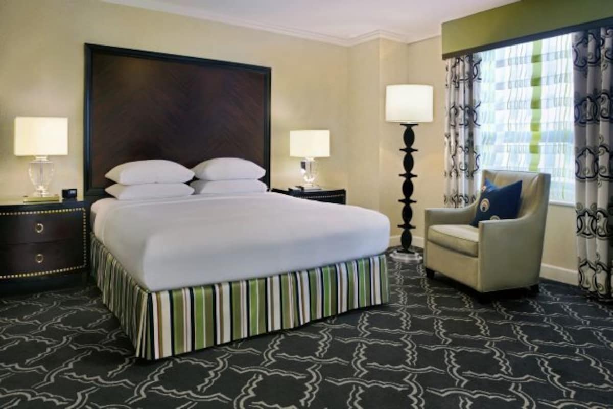 Quarto de hotel com uma cama grande, duas mesas de cabeceira com abajures, uma cadeira e uma janela com cortinas.