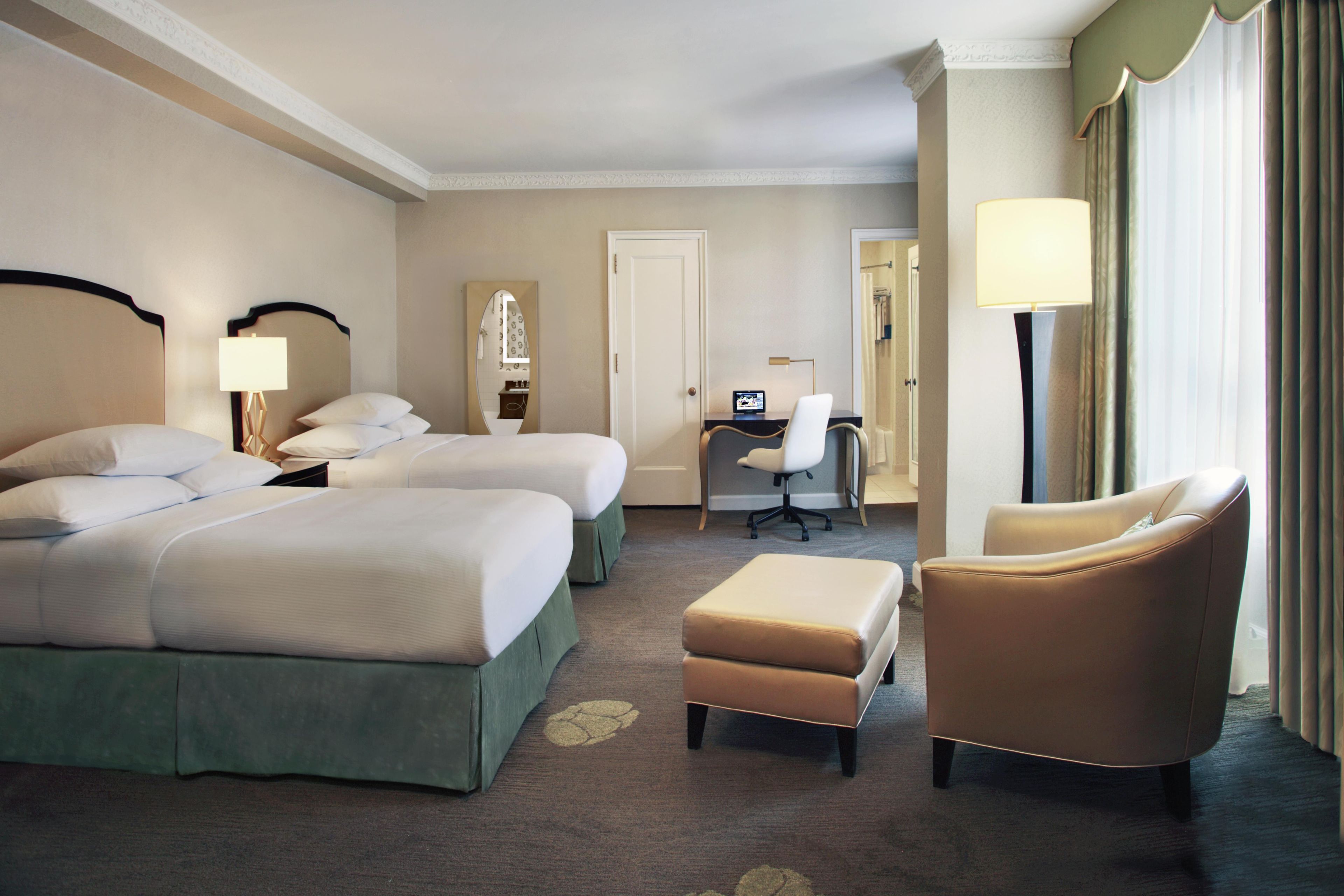 A hotel room with two beds, a desk, a chair, and a lamp.