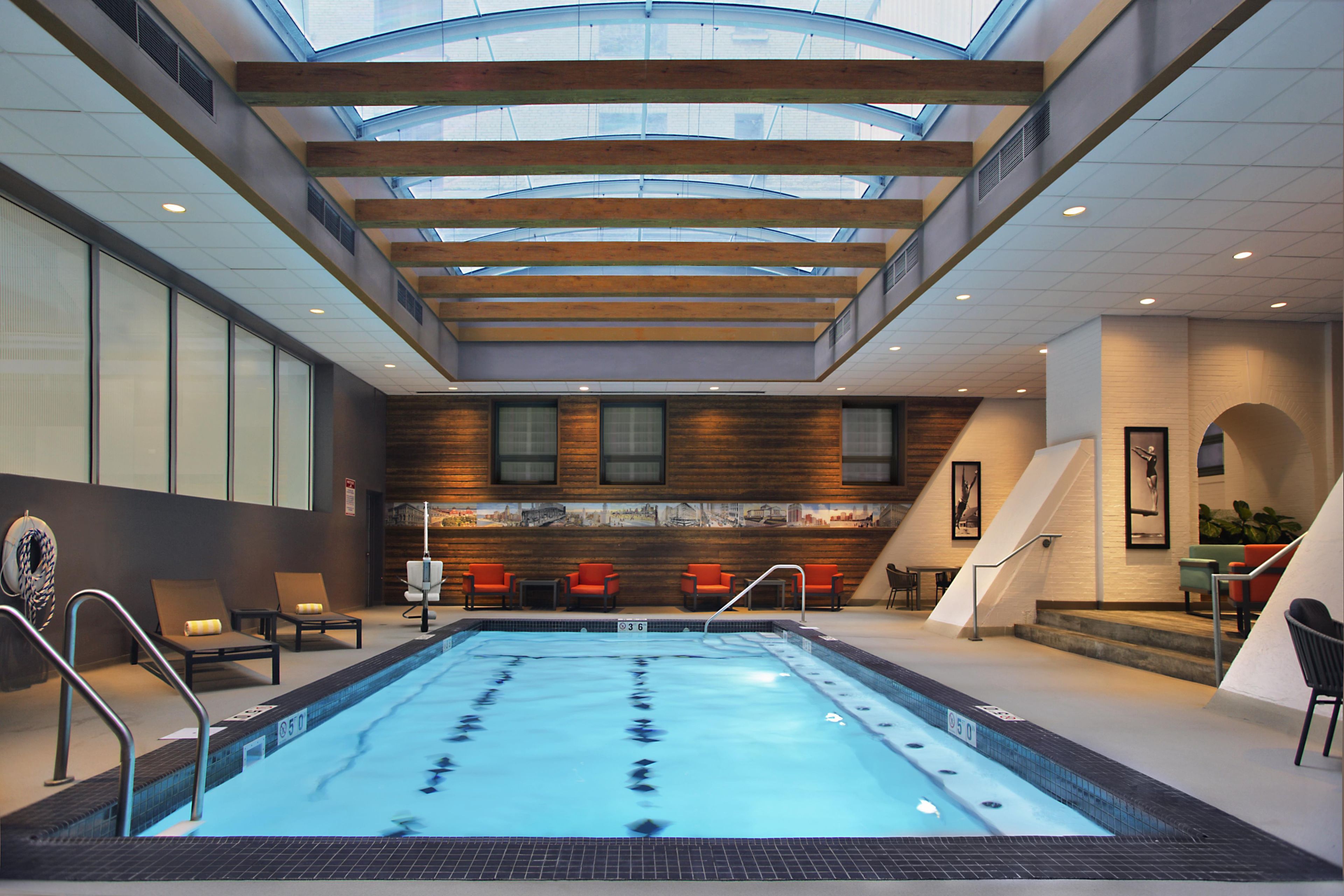 An indoor swimming pool with a skylight ceiling, wooden panel walls, and red chairs.