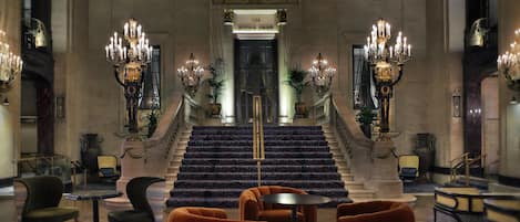 A grand lobby with a staircase, ornate chandeliers, and a clock on the wall.