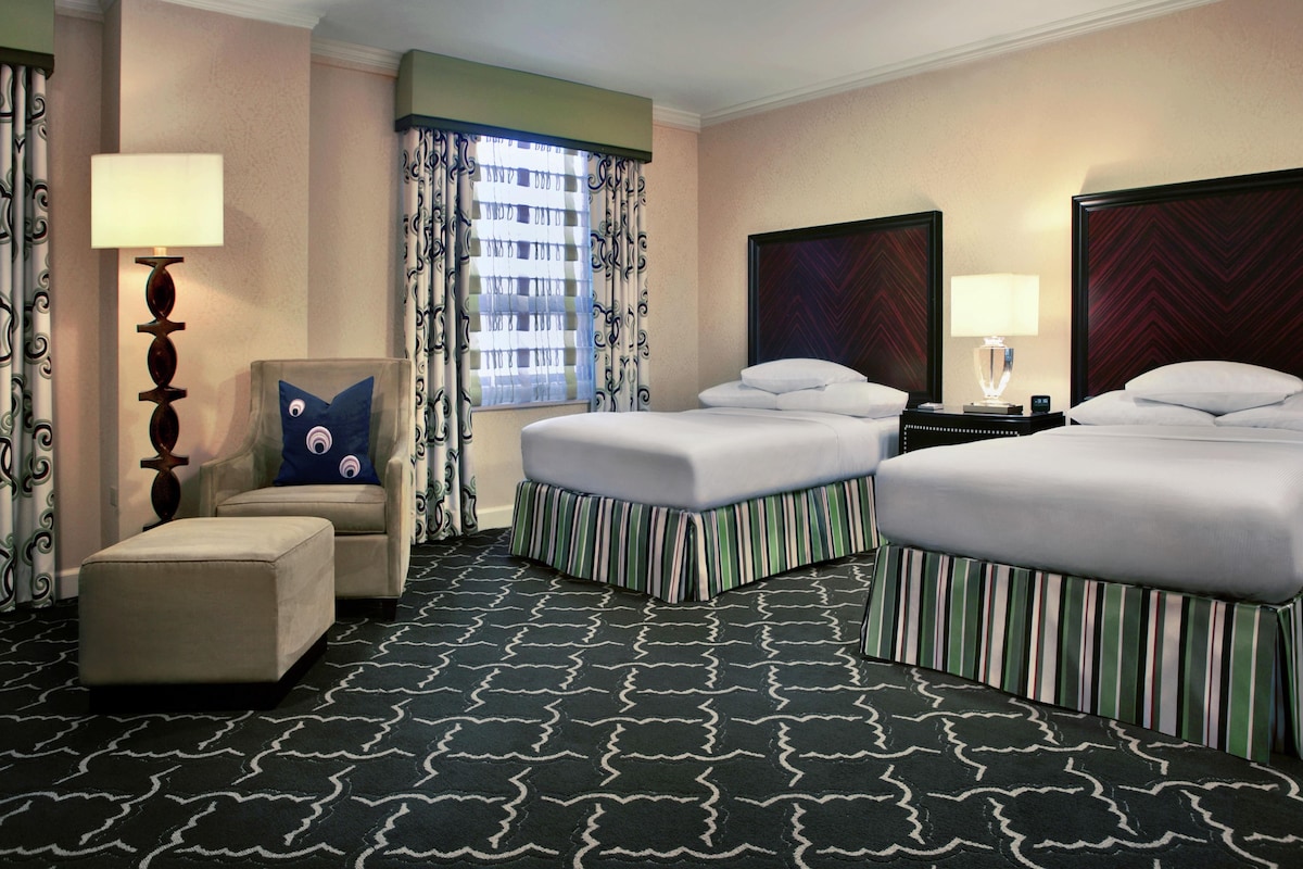 A hotel room with two beds, a chair, a desk, a lamp, and a window with curtains.