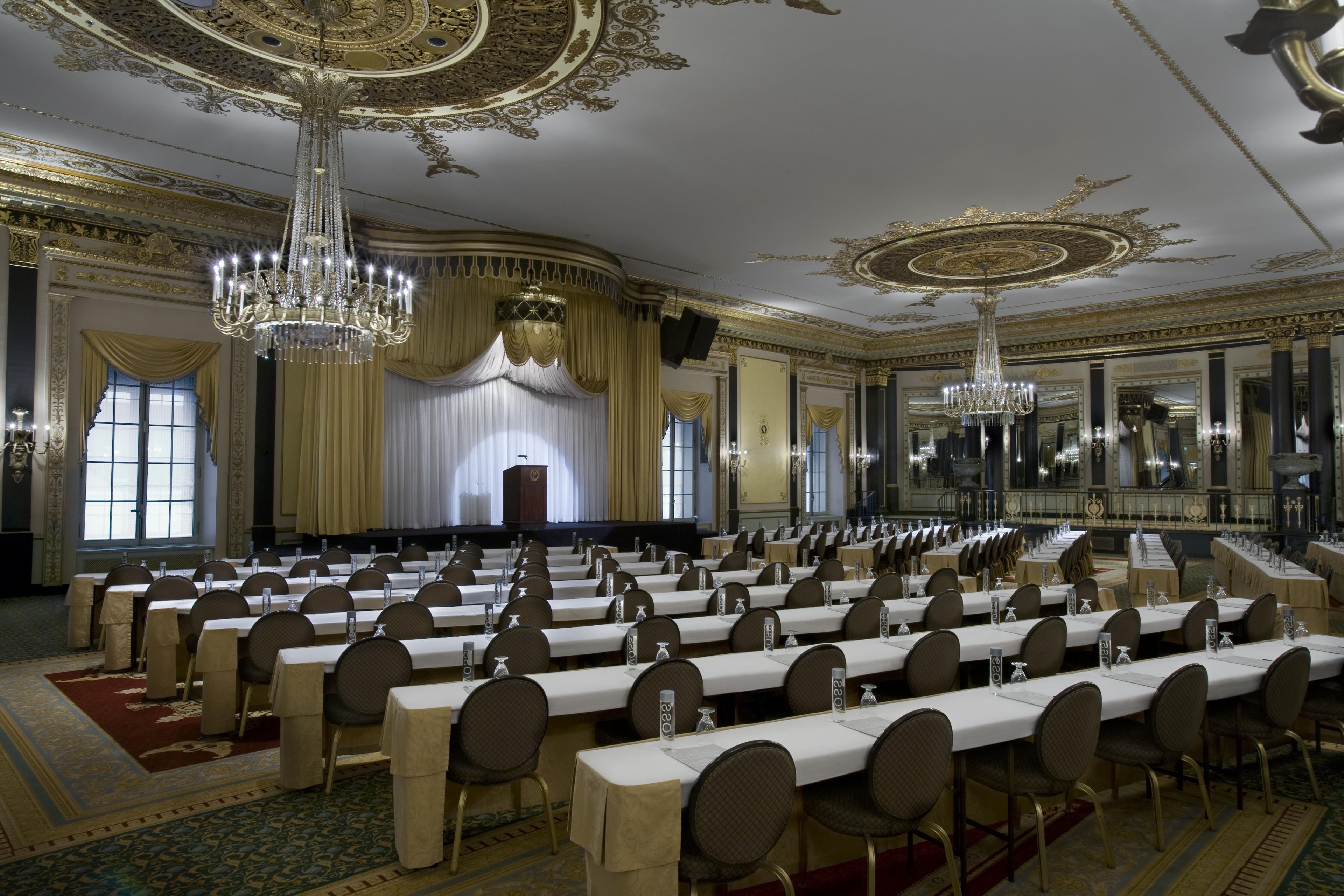 A grand hall with rows of tables and chairs, ornate chandeliers, and a stage with curtains.
