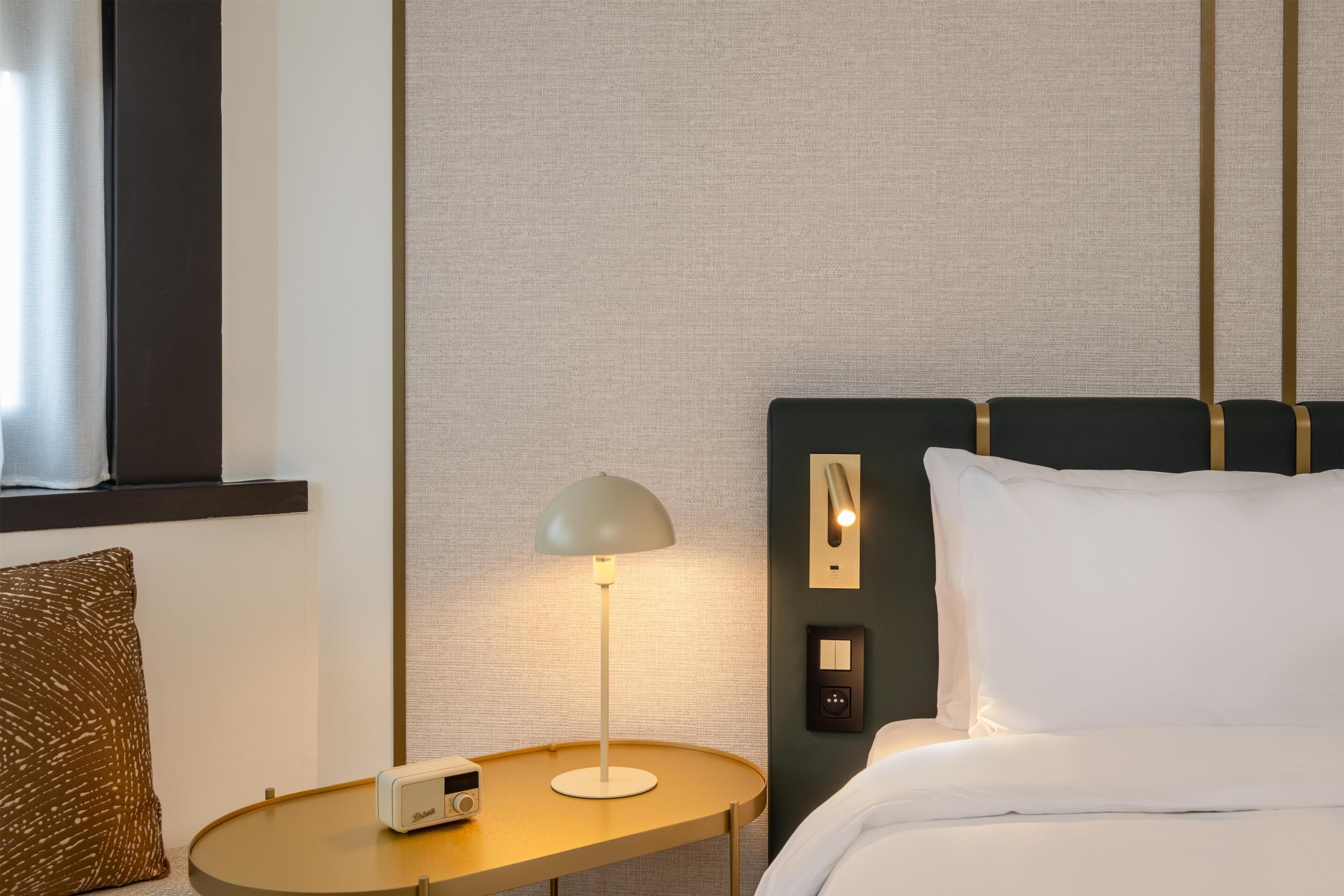 Quarto de hotel com cama, mesa de cabeceira com abajur, rádio e painel de controle fixado na parede.