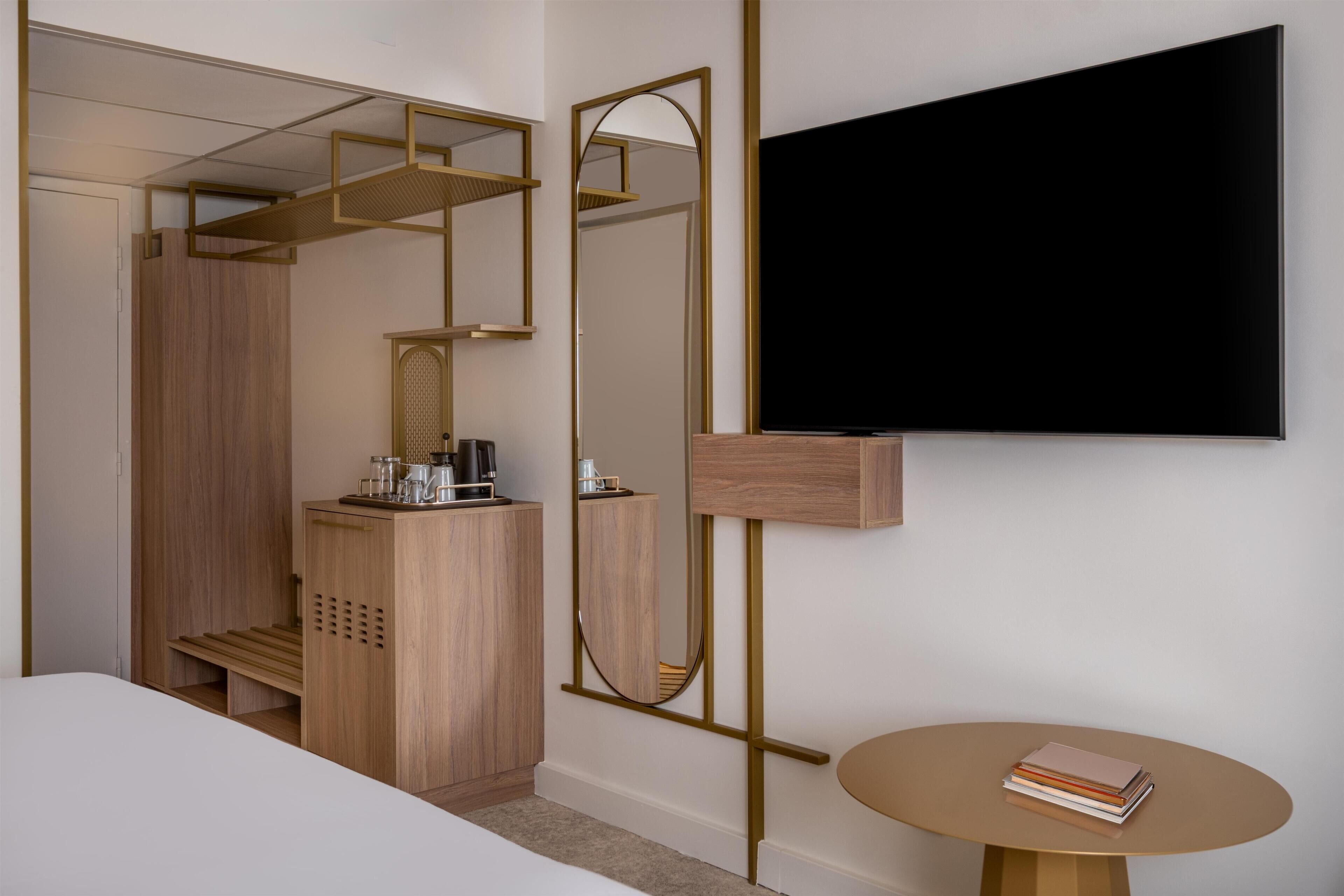 Una habitación moderna con una cama, un armario de madera, un escritorio con una cafetera, un televisor y una pequeña mesa redonda con libros.