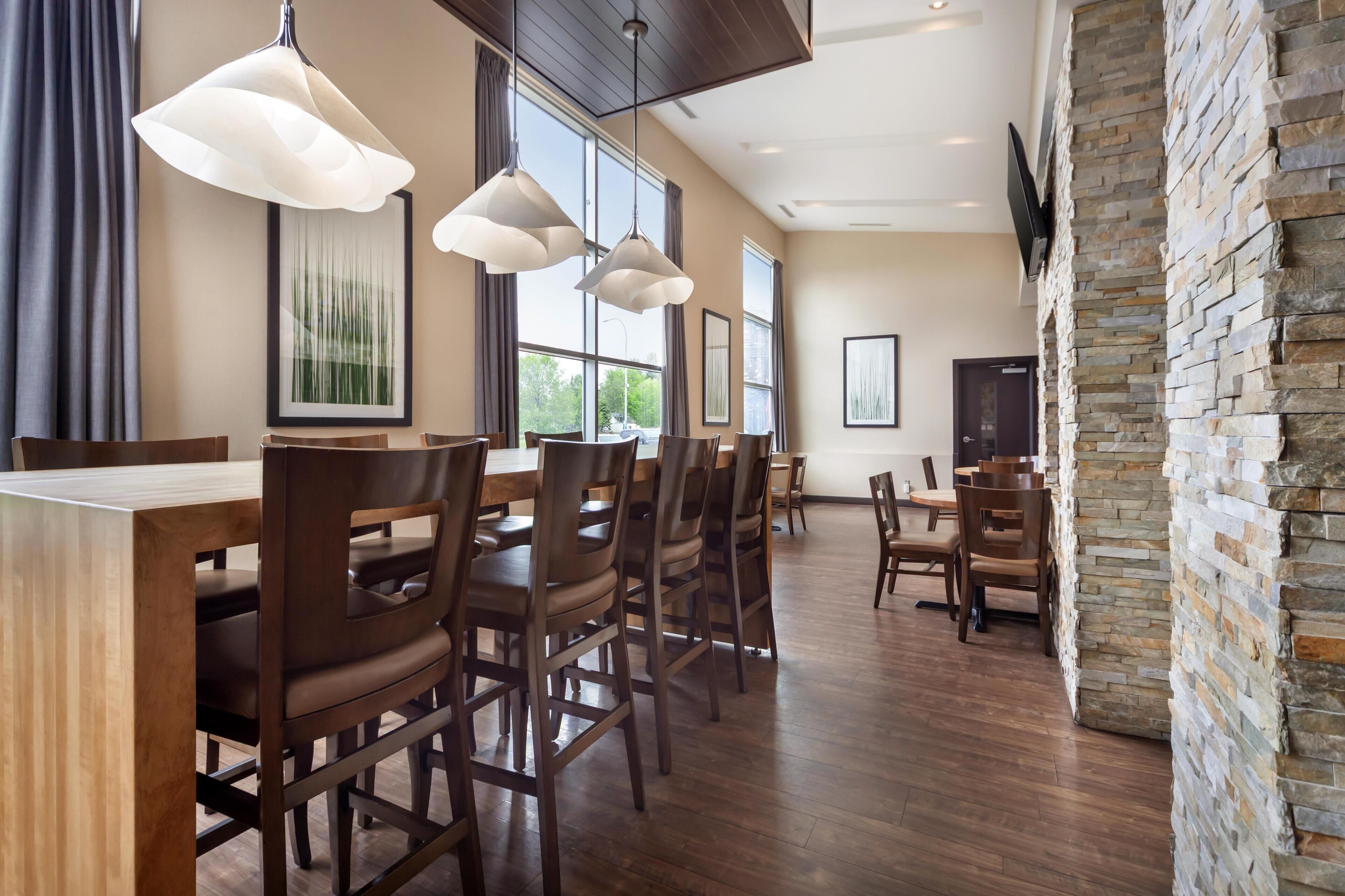 Comedor con mesas y sillas de madera, iluminación colgante, ventanas amplias y una pared decorativa de piedra.