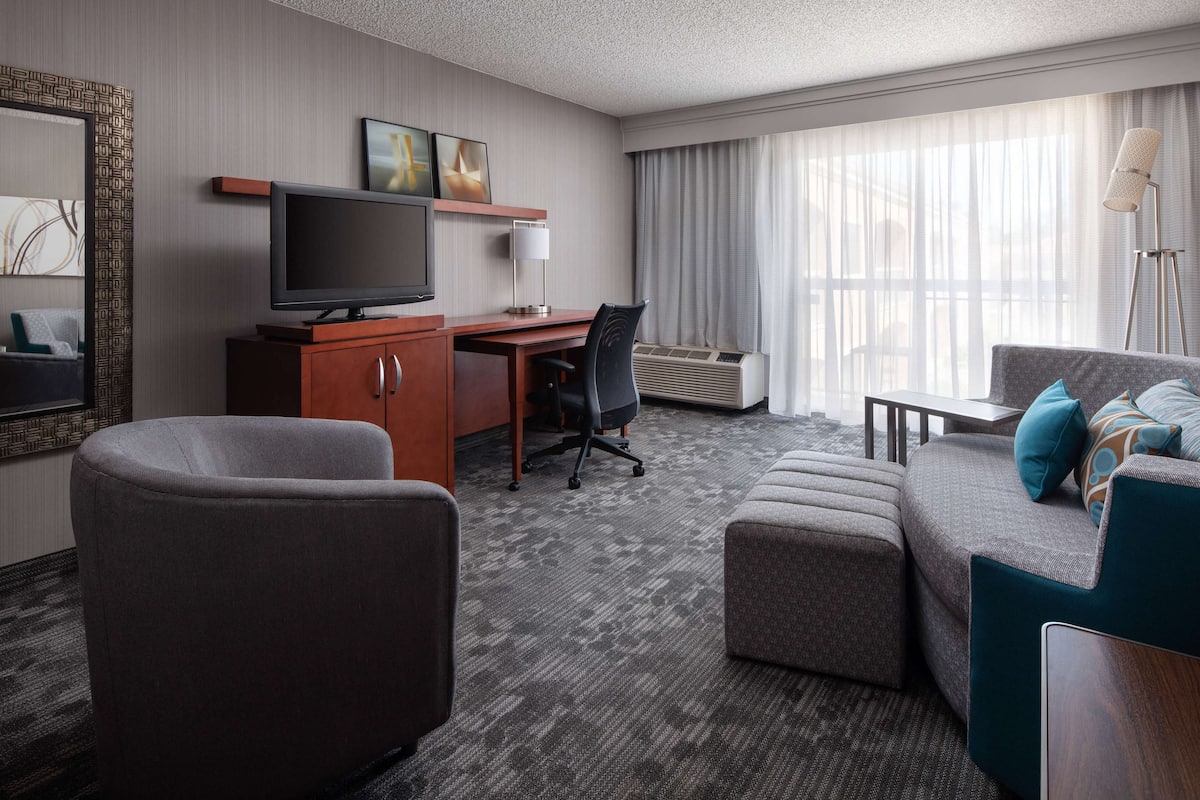 A hotel room with a large flat-screen TV, a desk with a chair, a sofa with cushions, and a standing lamp.