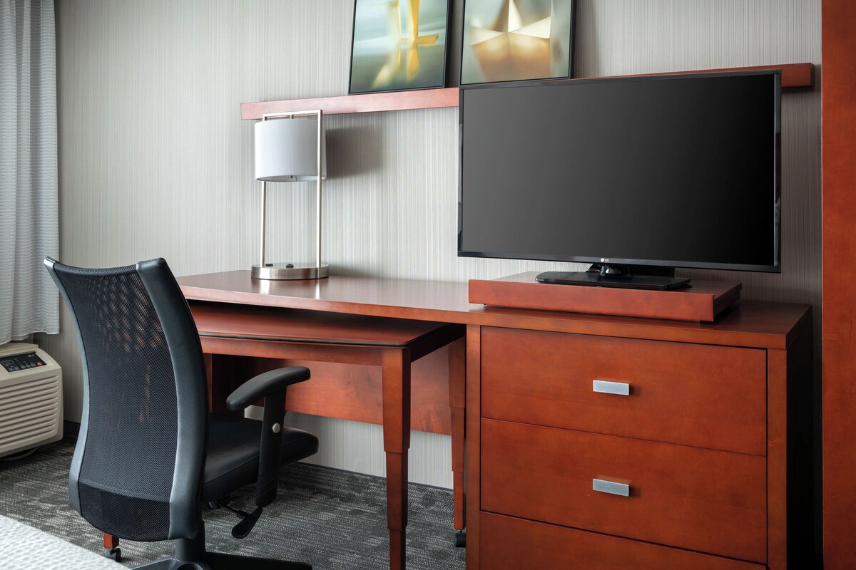 A hotel room with a wooden desk, a black office chair, a flat-screen TV, a lamp, and a small heater.