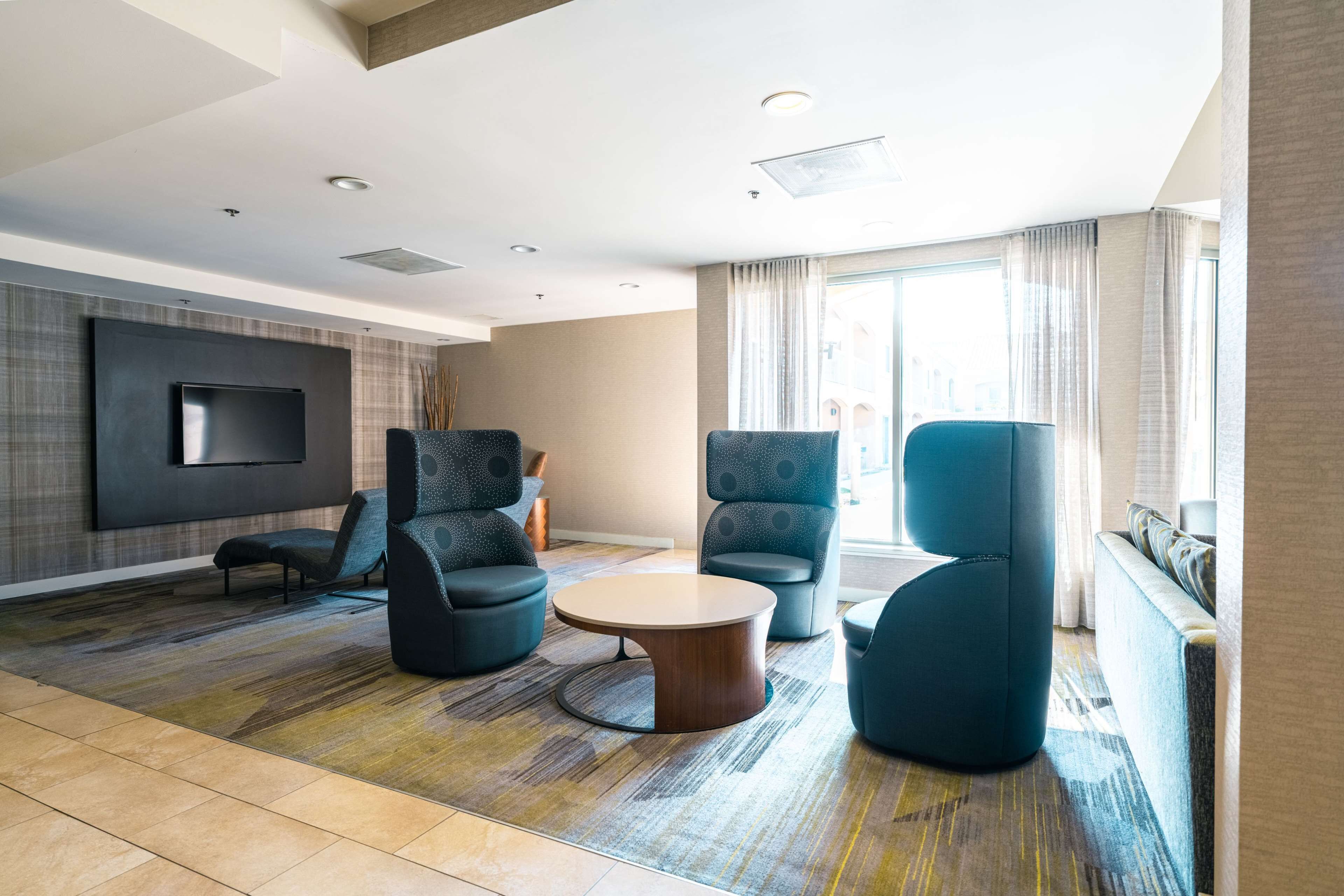 A modern hotel room with a large flat-screen TV mounted on the wall, a round coffee table, and several upholstered chairs.