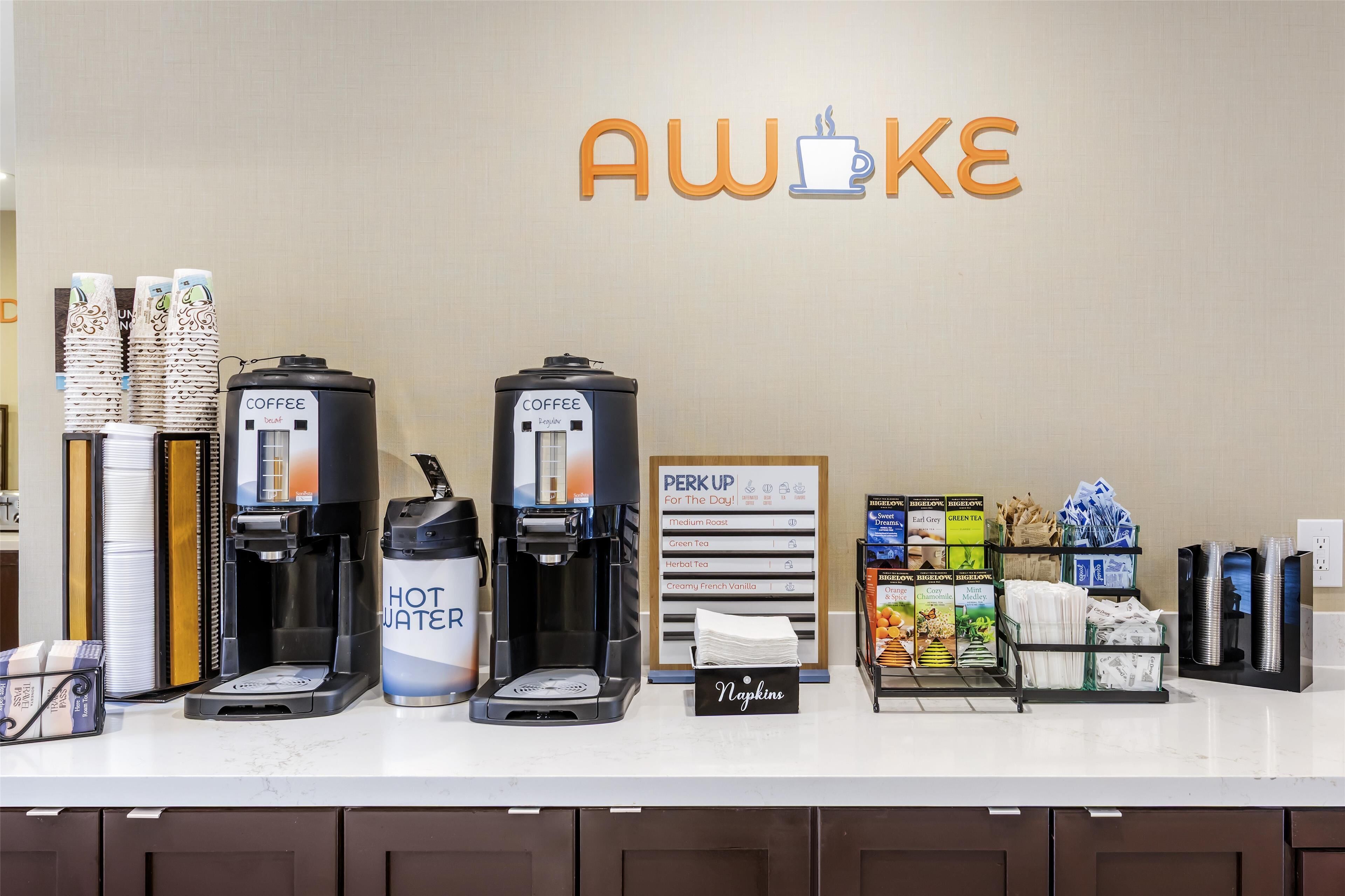 Un espace dédié au café, équipé de plusieurs machines à café, d’un distributeur d’eau et d’un panneau indiquant « Réveillez-vous et commencez bien la journée ! », avec différents choix de café.