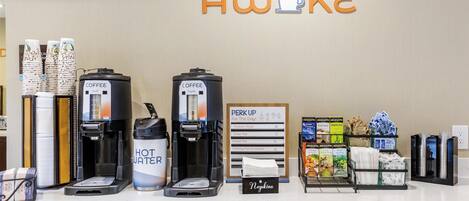 Un espace dédié au café, équipé de plusieurs machines à café, d’un distributeur d’eau et d’un panneau indiquant « Réveillez-vous et commencez bien la journée ! », avec différents choix de café.