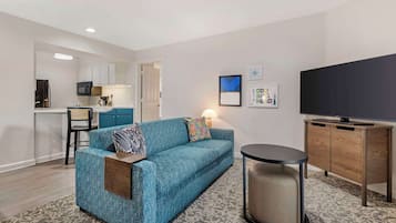 A modern living room with a blue sofa, a small round table, a television, and a kitchen in the background.