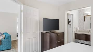 Un dormitorio con televisor sobre una cómoda, una cama, una silla azul y un baño con espejo y perchero.