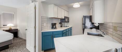 A compact hotel room with a kitchenette, a bed, a desk, and a chair.