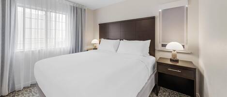 A hotel room with a large bed, a nightstand with a lamp, a mirror, and a window with sheer curtains.