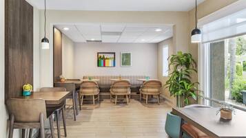 A modern dining area with wooden flooring, wicker chairs, and a bench. There are tables with potted plants and a large window providing a view of greenery.