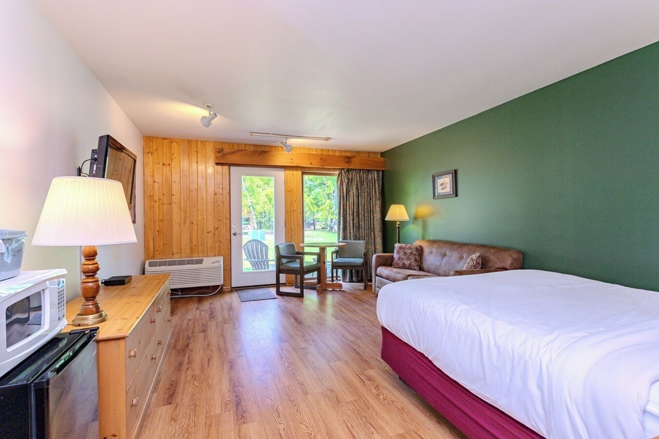 A hotel room with a bed, a desk with a lamp, a chair, a table, and a view of the outdoors.