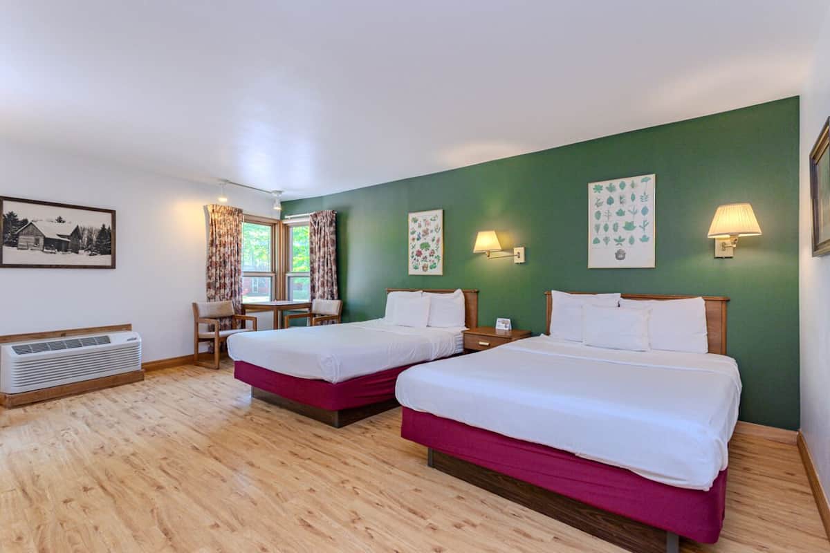 A hotel room with two beds, a desk, chairs, a window with curtains, wall-mounted lamps, and a heater.