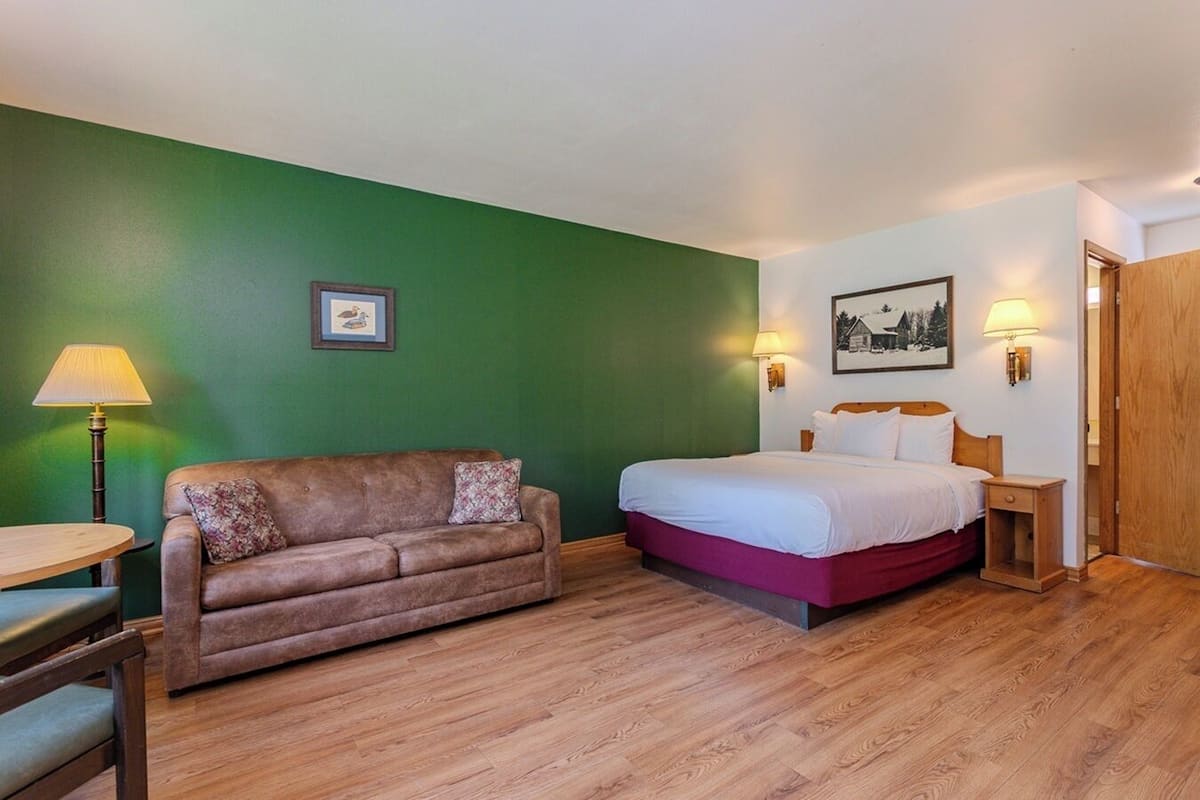 A hotel room with a green wall, a wooden floor, a brown sofa, a bed with white bedding, two bedside lamps, and a framed picture on the wall.
