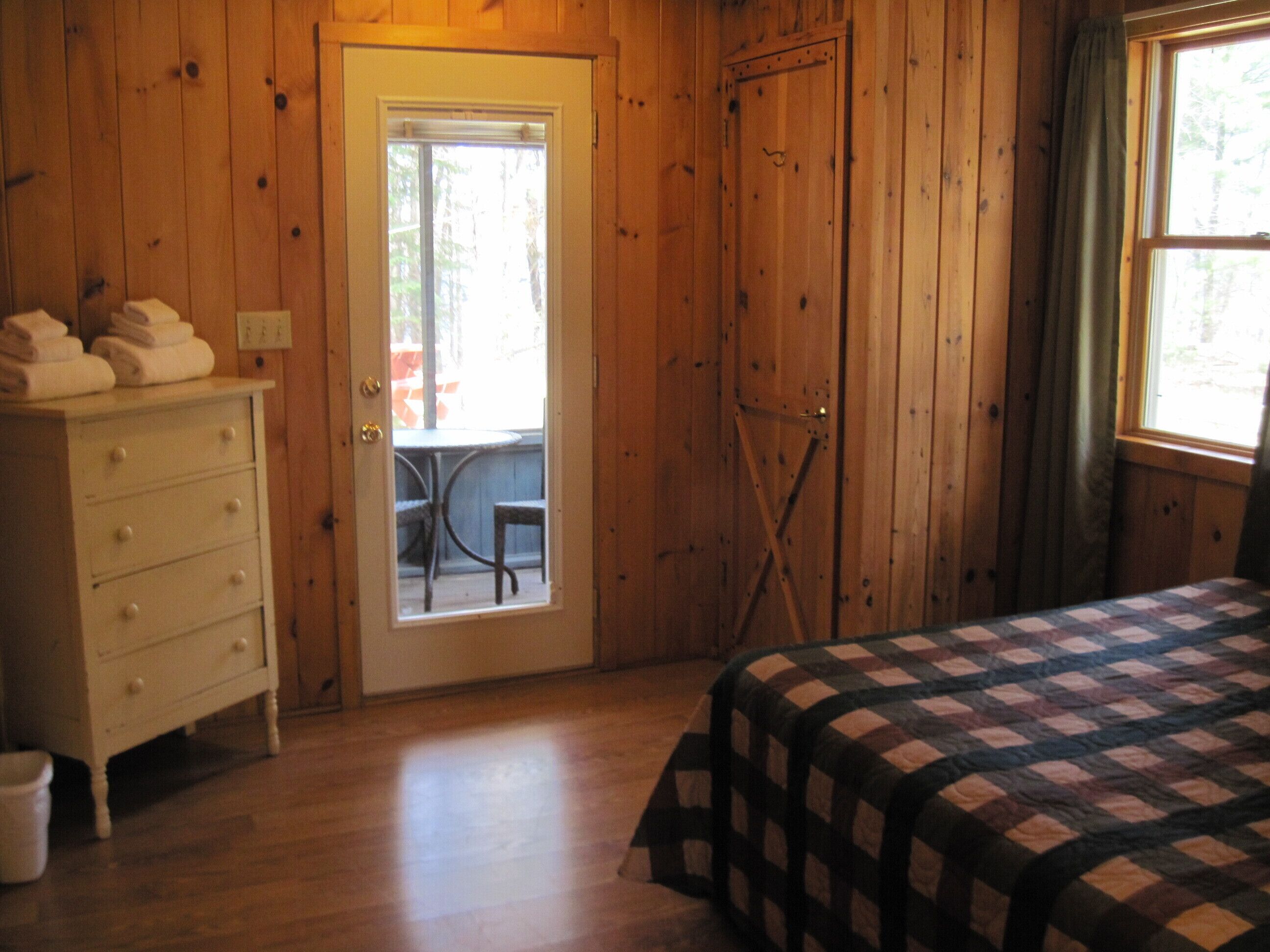 A wooden room with a bed, a dresser, and a door leading to an outdoor area with a table and chairs.