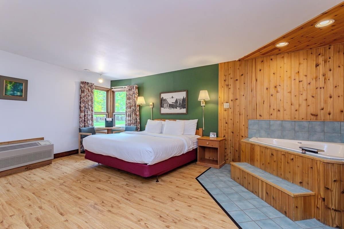 A hotel room with a bed, a desk, a chair, a window with curtains, a painting, a lamp, a wooden wall paneling, and a bathtub.