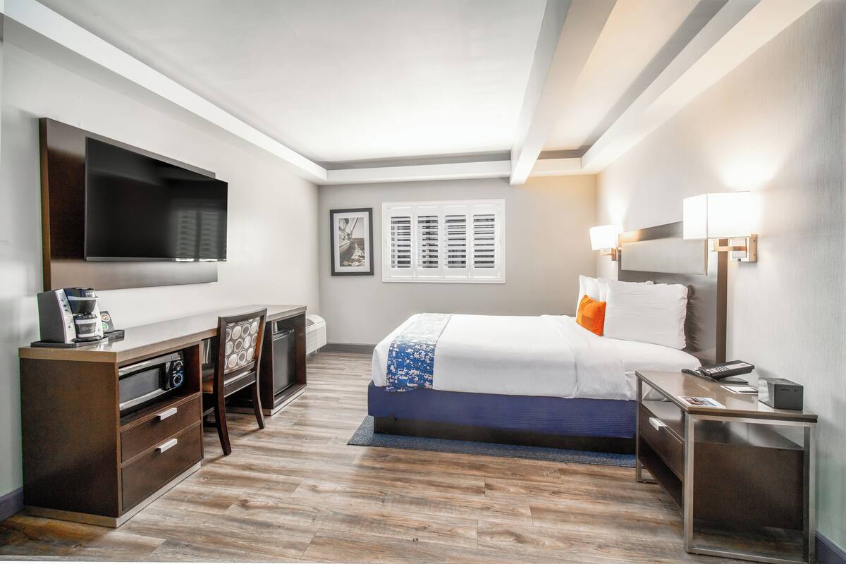 A hotel room with a bed, bedside tables, a desk with a chair, a flat-screen TV, a coffee maker, and a window with blinds.
