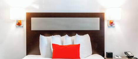 A hotel room with a large bed, a wooden headboard, two bedside lamps, a nightstand with a phone, and a red pillow.