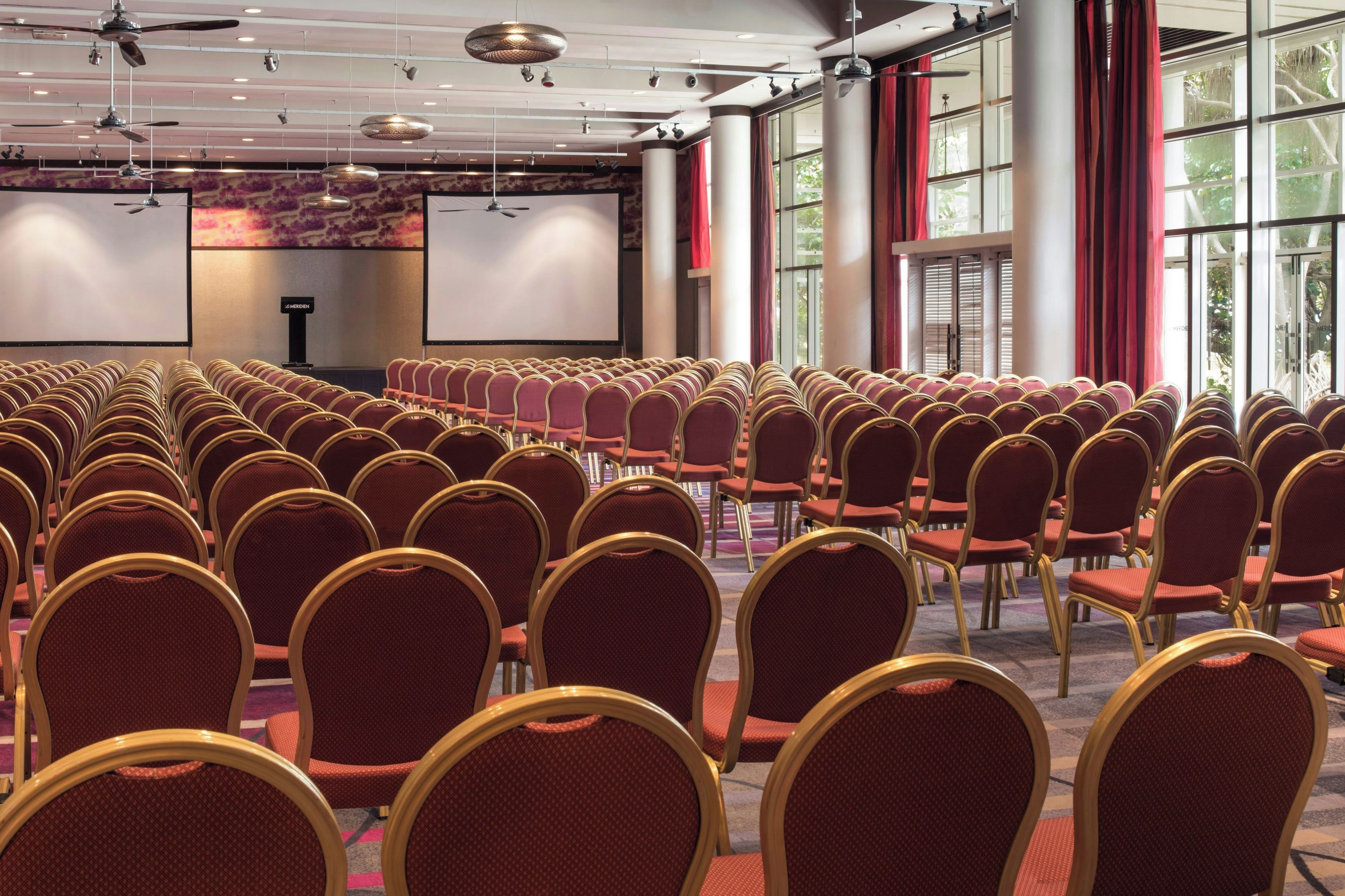 A conference room with rows of chairs facing a presentation screen, large windows, and ceiling fans.