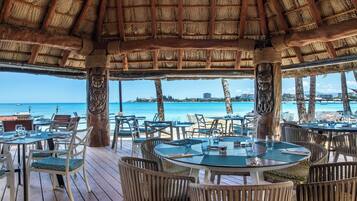 An outdoor restaurant with round tables and chairs, a thatched roof, and a view of the ocean.