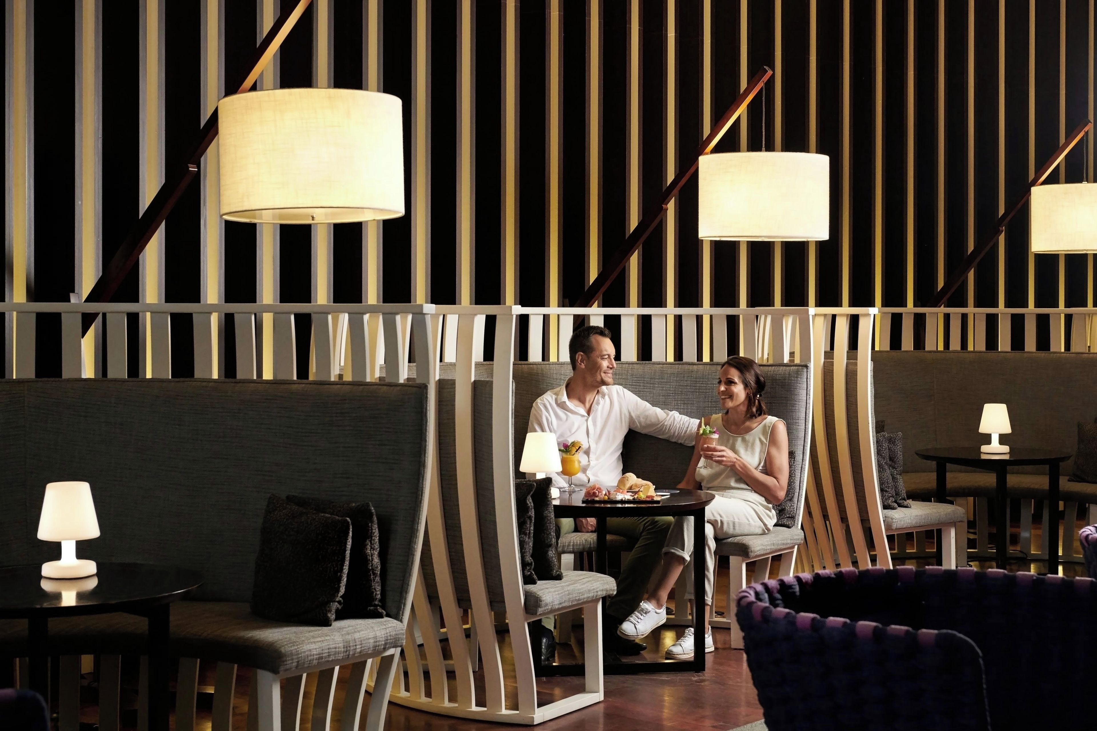 A couple seated on a sofa with a table between them, a lamp on a side table, and a patterned wall in the background.