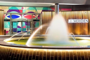 A hotel entrance with a fountain, a sign reading "Le MERIDIEN," and a colorful geometric wall design.