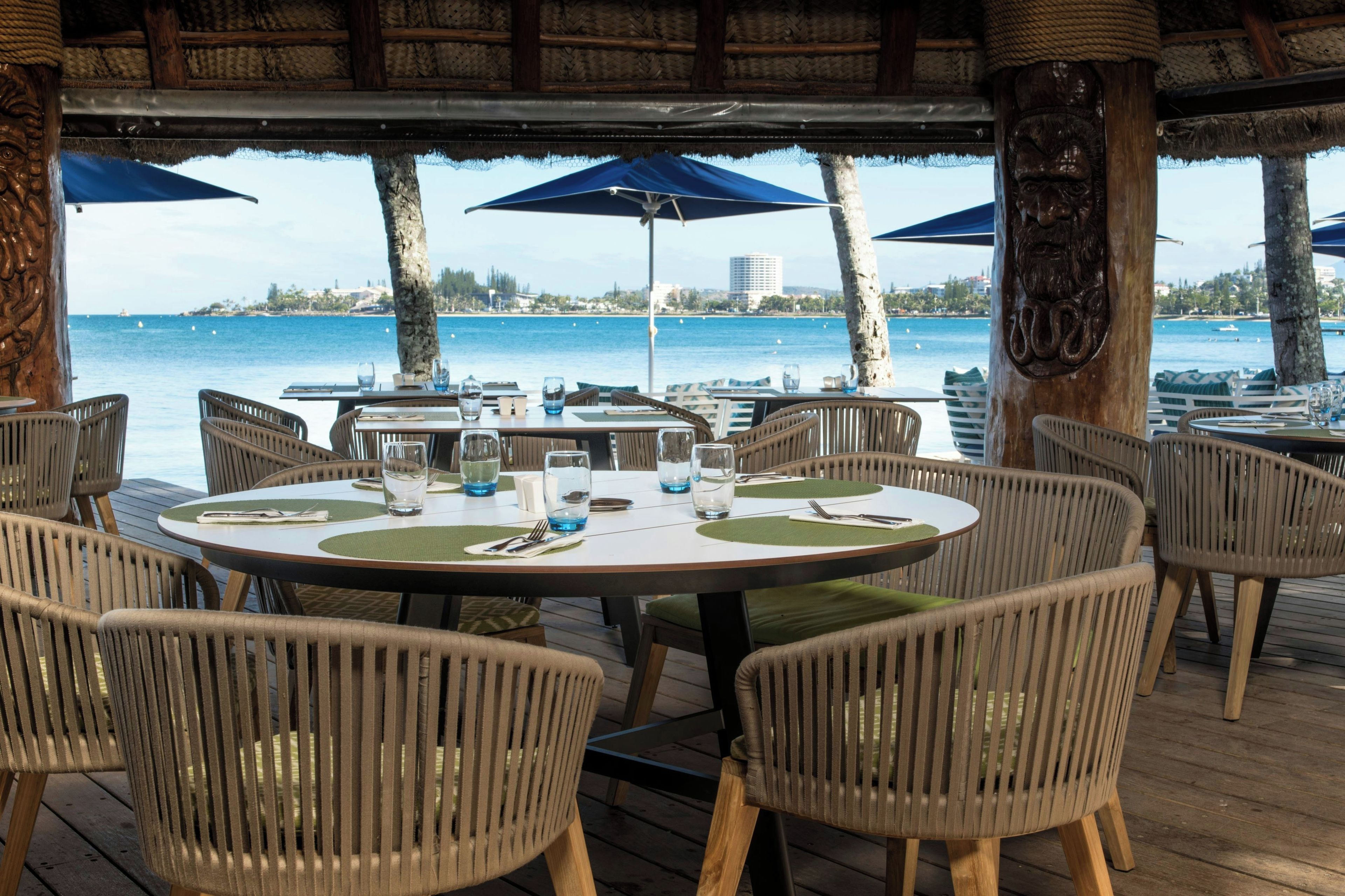An outdoor dining area with round tables and chairs, overlooking a body of water with a distant cityscape.